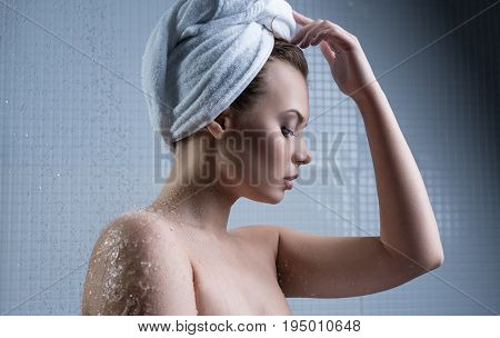 Young pretty girl her head wrapped in towel posing in shower surrounded by water splashes