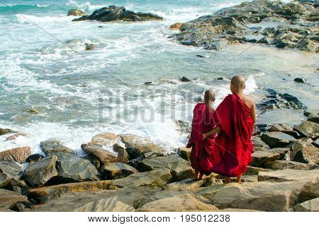 Two young Buddhist monks in red robes are standing at the shore of the ocean. They look at the waves. Southern province. Sri Lanka