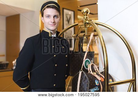 Smiling man in bellboy uniform standing with luggage cart and looking at the camera