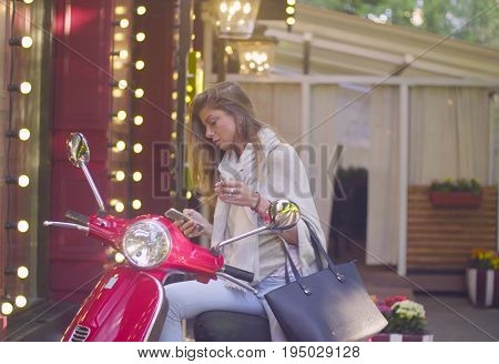 Portrait of young attractive woman sitting on the red scooter. She is searching something in smartphone