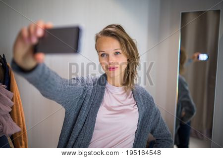 Young woman shopping in a fashion store, trying on some clothes she likes  - taking a selfie picture with her smartphone in the cabin (color toned image; shallow DOF)