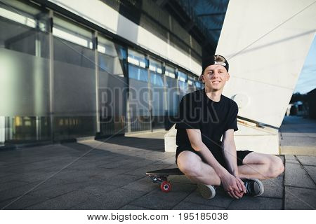 A smiling teenager skater sits on a blackboard on the background of modern architecture. A skater resting on a board for skiing.