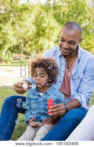 Smiling father and son playing with soap bubbles at park. Little african boy blowing soap bubble in park with dad. Black father and son playing together and sitting on grass.