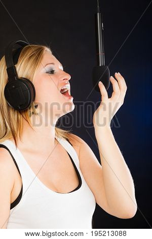 Music passion stage-fright concept. Blonde young woman singing to microphone and wearing big headphones on her head performing songs in studio.