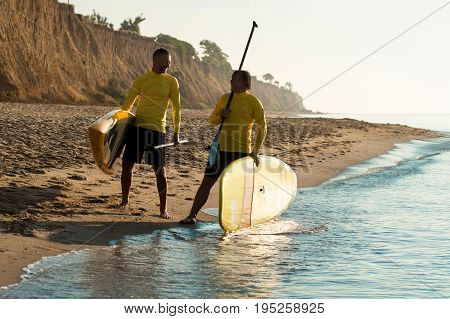 two sports man walking along the seashore with a surf board and paddle
