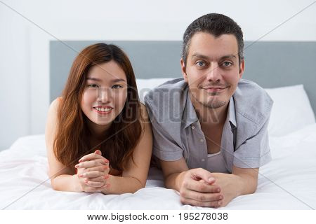 Positive intercultural couple lying on soft duvet in bedroom and looking at camera. Portrait of happy lovers relaxing together in bed. Honeymoon concept