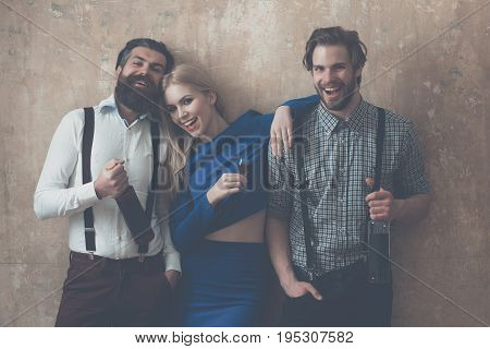 Woman holding glass of liqueur and men with wine bottles. Happy friends with alcoholic drinks on beige background. Alcohol and appetizer. Friendship and convives. Unhealthy lifestyle and bad habits