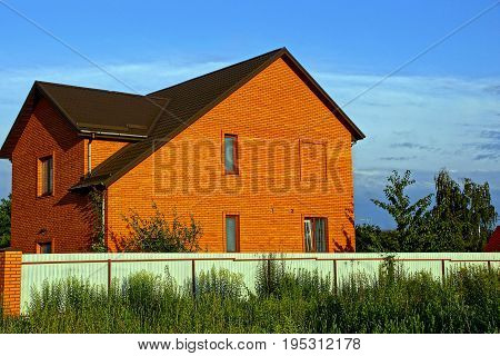 The facade of a brown brick house outside a fence in the garden and greenery