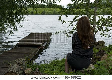 girl with long hair female one 1 sits on the banks of the river a wooden platform trees grass water