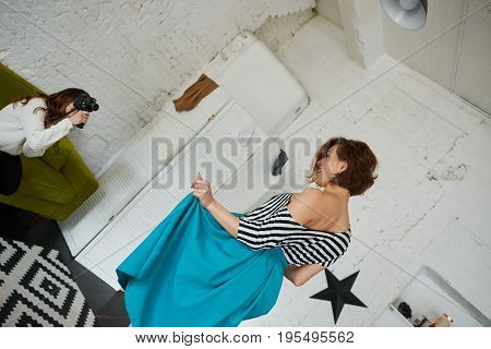 Two female friends having fun at home: woman with camera taking picture of girl with short hair who posing in striped top and turquoise skirt. People lifestyle friendship and technology concept