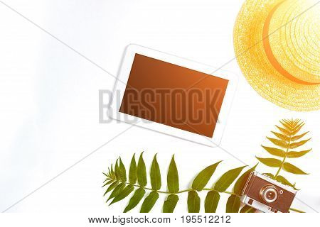 Straw hat with green leaves, tablet and old camera on white background, Summer background. Top view. Copy space. Still life. Flat lay. Sun flare