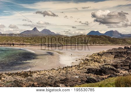 Assynt Peninsula Scotland - June 7 2012: Achnahaird Beach is sandy with gentle Atlantic Ocean waves out of Enard Bay. Lone walker far on beach. Mountainous background. Rocky foreground. Heavy clouds