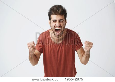 Happy positive excited young man clenching fists and screaming, wearing casual t-shirt glad to hear good news, celebrating his victory or success. Life achievement, goals and happiness concept.
