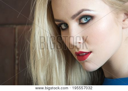 Girl with red lips on adorable face. Pretty woman fashionable model with blue eyes bright makeup and long blond hair posing on brown wall. Fashion make up visage and cosmetics. Beauty salon