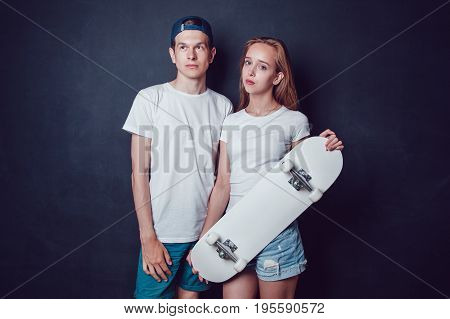 Young skater boy and girl with skateboard in white T-shirts standing on black background.