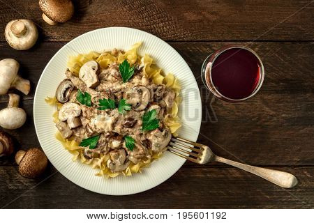 Mushroom beef stroganoff, served with the typical egg pasta, with cremini and champignons, with a glass of red wine, and a fork, shot from above on a dark rustic texture with a place for text
