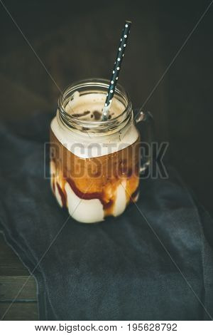 Iced caramel macciato coffee with milk in glass jar with straw over dark rustic wooden table background, selective focus, copy space. Cold sweet refreshing summer drink concept