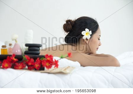 Asian Woman Lying Down Relaxation On White Bed In Spa Salon With Flowers, Oil And Hot Stone. Beautif