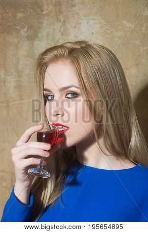 Woman drinking glass of red liqueur. Girl with blond long hair make up in blue dress on beige wall. Alcohol appetizer bad habits addictive and convive. Unhealthy lifestyle