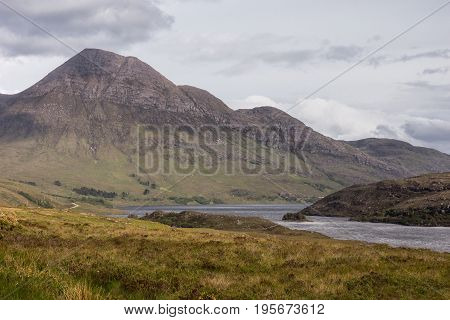 Assynt Peninsula Scotland - June 7 2012: Brown Mountain range along Loch Lurgainn under silver sky with gray clouds. Green vegetation on foothills and dry foreground.