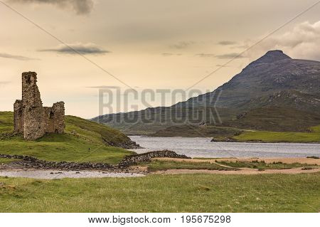 Assynt Peninsula Scotland - June 7 2012: Ruins of Castle Ardvreck at Loch Assynt with mountain in back under yellowish twilight sky. Green ground vegetation.