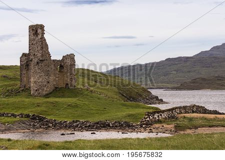 Assynt Peninsula Scotland - June 7 2012: Brownish Ruins of Castle Ardvreck from across Loch Assynt. Green ground vegetation. Brown mountain slope. Silver sky.