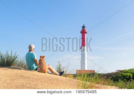 Man and his dog sitting on dune and looking to Lighthouse red and white at the French west beach