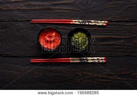 Pieces of traditional japanese tablewear. Red chopsticks with hierogliphs, small black bowls with ginger and wasabi on dark wooden table. Top view, flat lay, copy space, background