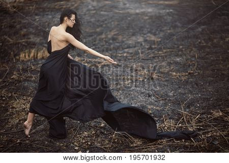 Sad girl standing on the burned-out land. Concept of environmental disaster. Global problems with ecology. Young attractive multi-racial Asian Caucasian woman in black dress on nature