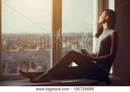 Resting and thinking woman with closed eyes. Calm girl with cup of tea or coffee sitting on the window-sill at home. Side view. Young attractive multi-racial Asian Caucasian lady in casual clothes resting indoors