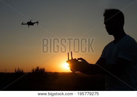 Silhouette of a man piloting drone in the air with a remote controller in his hands on sunset. Pilot takes aerial photos and videos with quadcopter from above. Copter on background
