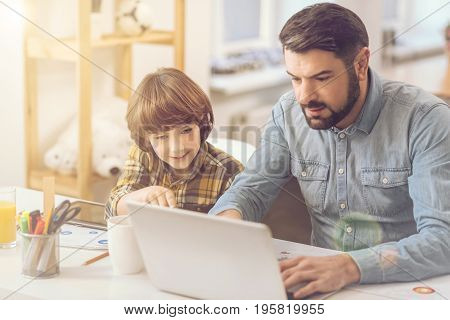 Helping with a home task. Handsome nice caring father sitting at the laptop and typing while helping his son with home task