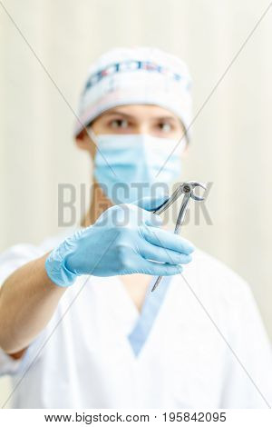 Portrait of handsome male dentist with stainless steel dental tongs or pliers for extraction of lower teeth in the dental clinic. Selective focus on dental tongs
