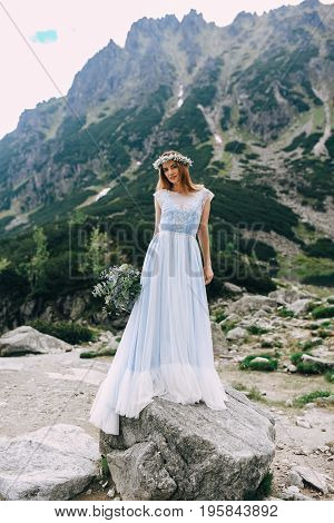 the bride in a beautiful blue dress and a wreath of white flowers holding a bouquet of purple flowers and eucalyptus overlooking the green mountains