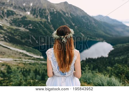 the bride in a beautiful blue dress with a deep neckline at the back with a wreath on her head of white flowers stands on a background of mountains and lakes