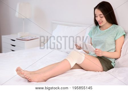 Beautiful young woman with tablet computer and bandaged leg sitting on bed at home