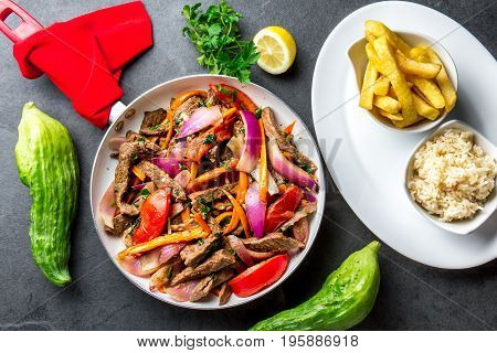 Peruvian dish Lomo saltado - beef tenderloin with purple onion, yellow chili, tomatoes in pan, fried potatoes french fries and rice on slate background. Tot view