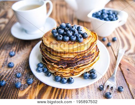 Pancakes healthy breakfast with blueberries, bog whortleberry, cup of green tea, cup of blueberries and teapot on brown wooden background