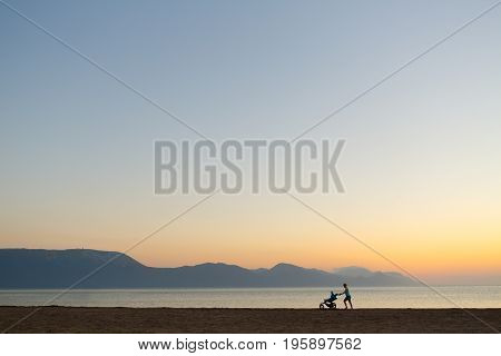 Silhouette of mother with stroller enjoying motherhood at sunset landscape. Walking or jogging woman with pram on a beach at sunset. Beautiful inspirational mountains landscape.