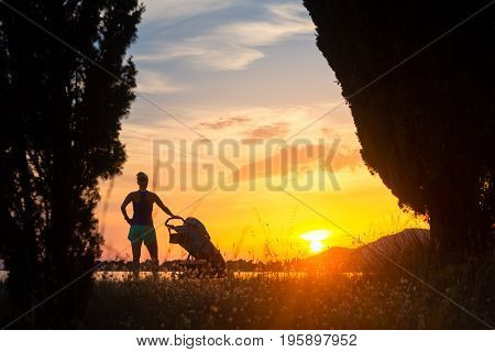 Silhouette of mother with child in stroller enjoying motherhood at sunset landscape. Walking or jogging woman with pram on a beach at sunset. Family relaxing at beautiful inspirational mountains landscape.