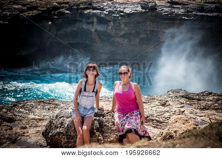 Two young and sexy women on the rocks near the wild ocean. Storm, huge waves coming and splashing. Tropical island Nusa Lembongan. The name of the place is Devils Tear.