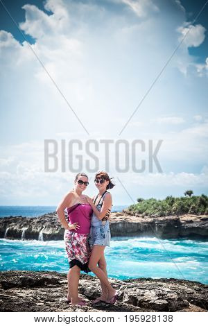 Two young and sexy women on the rocks near the wild ocean. Storm, huge waves coming and splashing. Tropical island Nusa Lembongan. The name of the place is Devils Tear.