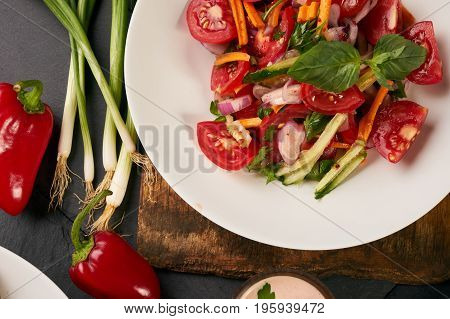 Fresh summer low-calories vegetable salad with tomatoes cucumbers carrots onions fresh bell peppers and garlic on wooden board over the dark stone background. Heathy vegetarian food and detox concept