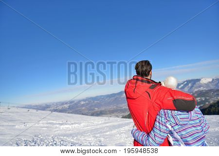 Rear view of a loving couple in fur hood jackets looking at snowed mountain range.