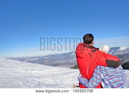 Rear view of a loving couple in fur hood jackets looking at snowed mountain range.