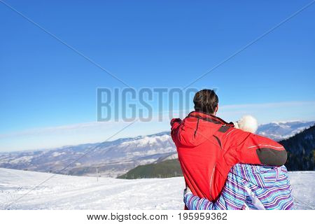 Rear view of a loving couple in fur hood jackets looking at snowed mountain range.