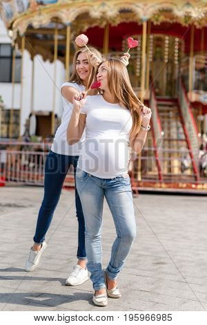 Two pretty women in amusement park having fun and eating sweets, happy family. Samesex lesbian family, pregnant couple