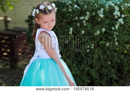 little girl is shy and looks down сute small girl dressed in blue and white dress with a wreath of artificial flowers on her head child in a festive dress on a nature background