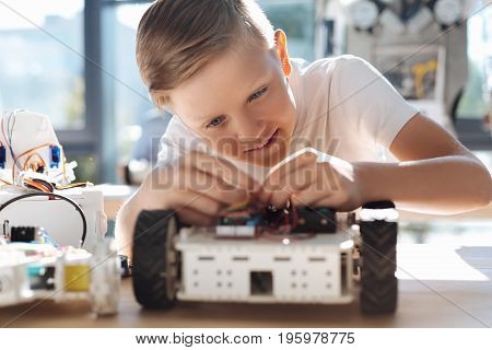 Diligent student. Pleasant fair-haired boy sitting at the robotics class and thoroughly fixing wires in his robotic car, being totally immersed into this process