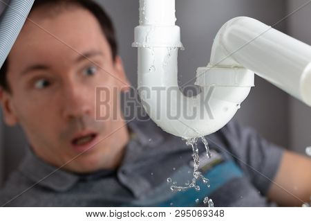 Shocked Young Man Looking At Sink Pipe Leakage In Kitchen